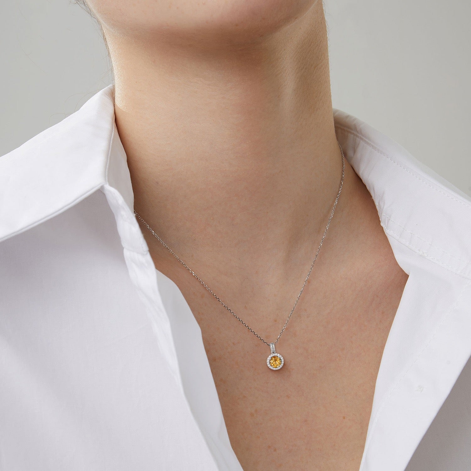 Gros plan d'un collier en argent S925 avec une citrine ronde et lumineuse au centre, sertie dans un halo de petites pierres scintillantes, porté par une femme sur une chemise blanche.