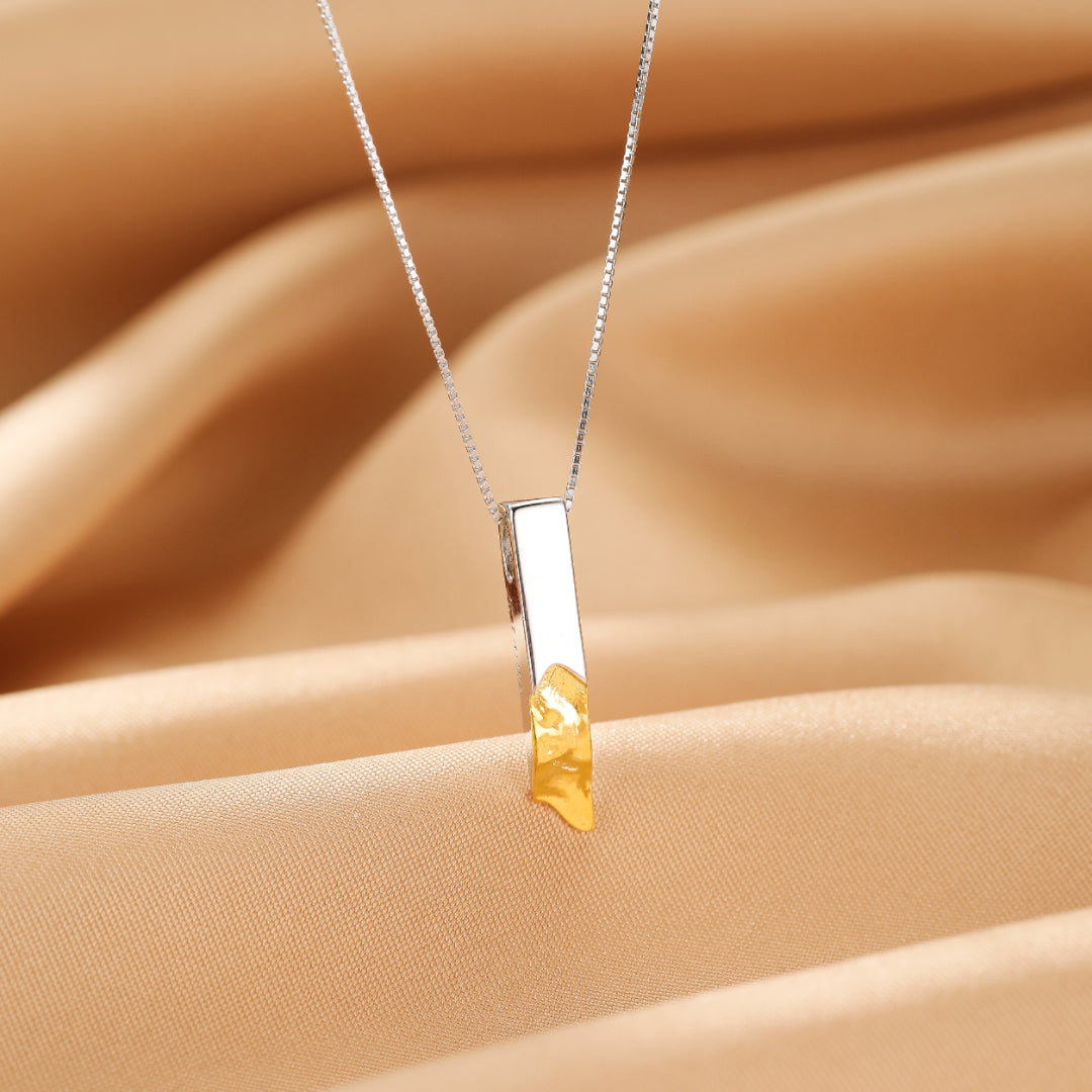 Collier pendentif barre argentée avec une touche dorée texturée, sur fond satiné doré.
