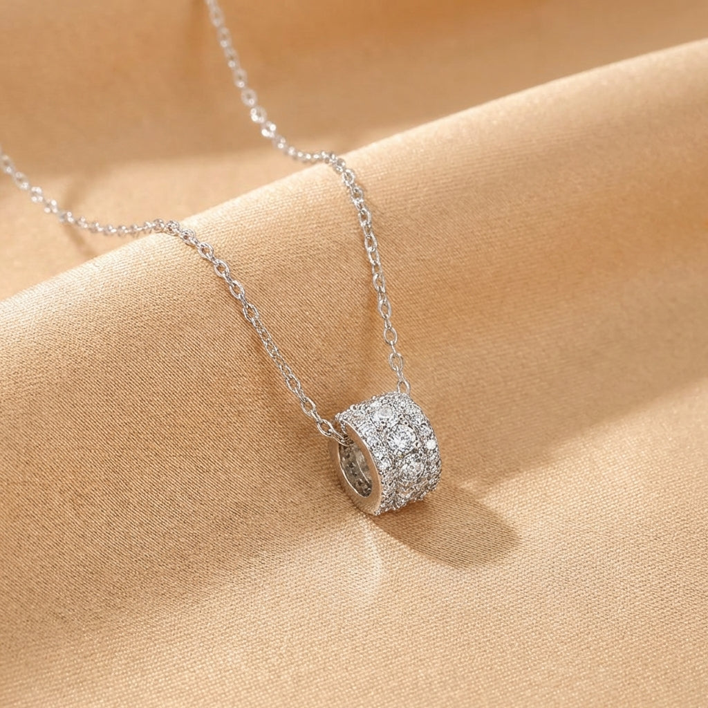 Collier en argent avec un pendentif cylindrique serti de cristaux d'imitation scintillants, posé sur un tissu satiné couleur sable.