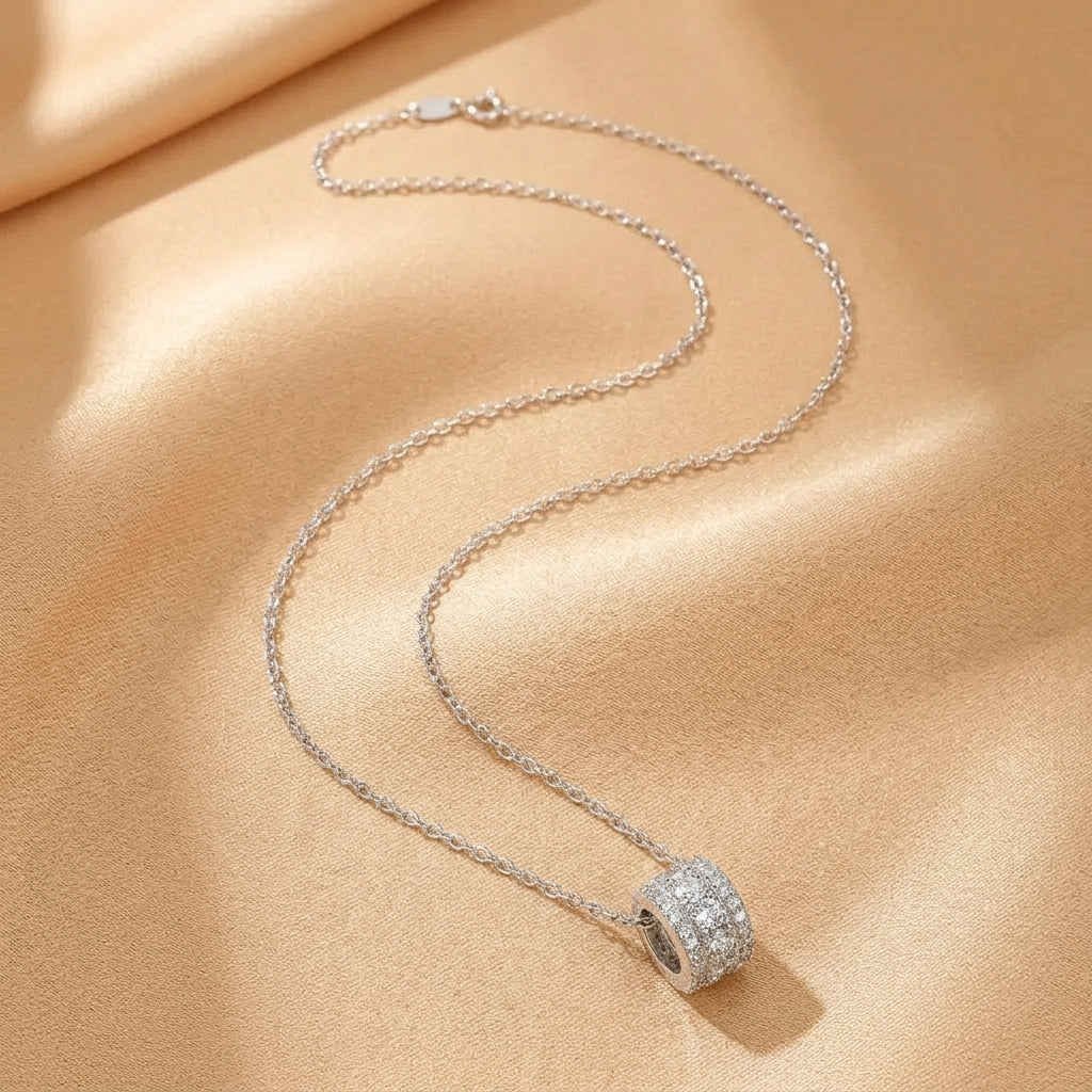 Collier argenté avec un pendentif cylindrique serti de cristaux brillants sur un fond satiné beige.