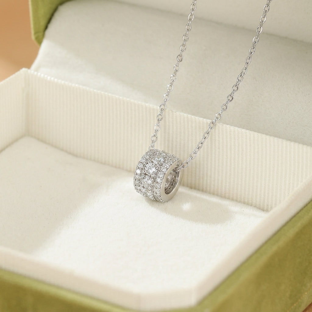 Un collier en argent avec un pendentif cylindrique serti de cristaux étincelants, présenté dans une boîte à bijoux verte.