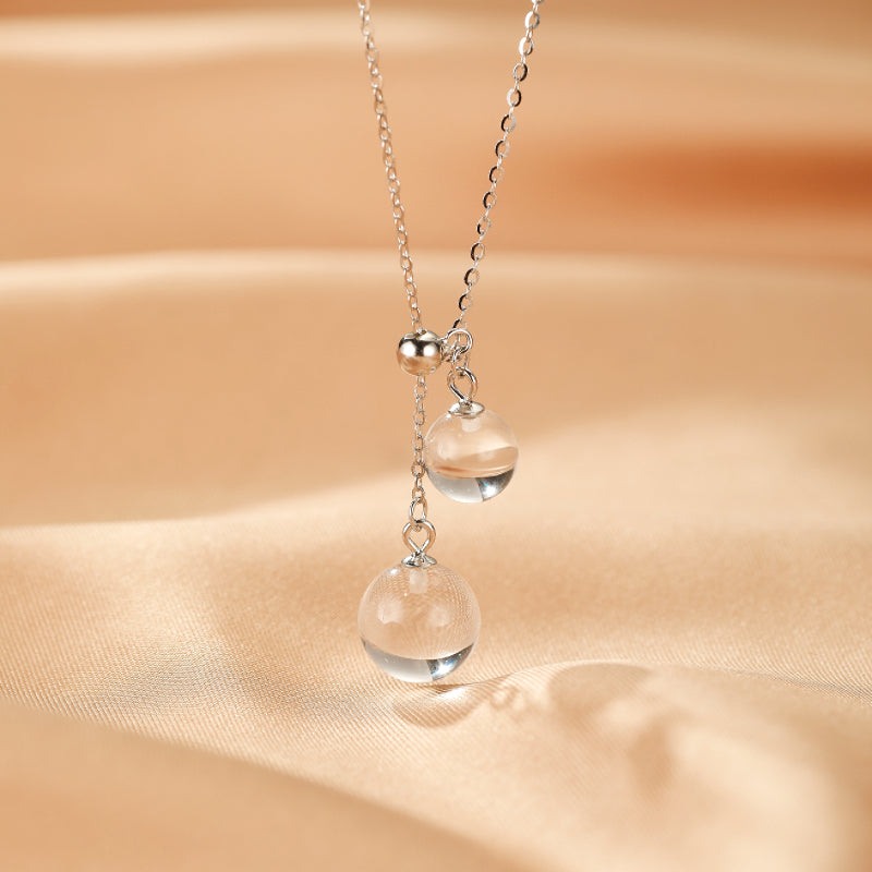 Collier en argent avec deux pendentifs sphériques en cristal de roche, suspendus à des longueurs différentes, sur un fond de satin beige.