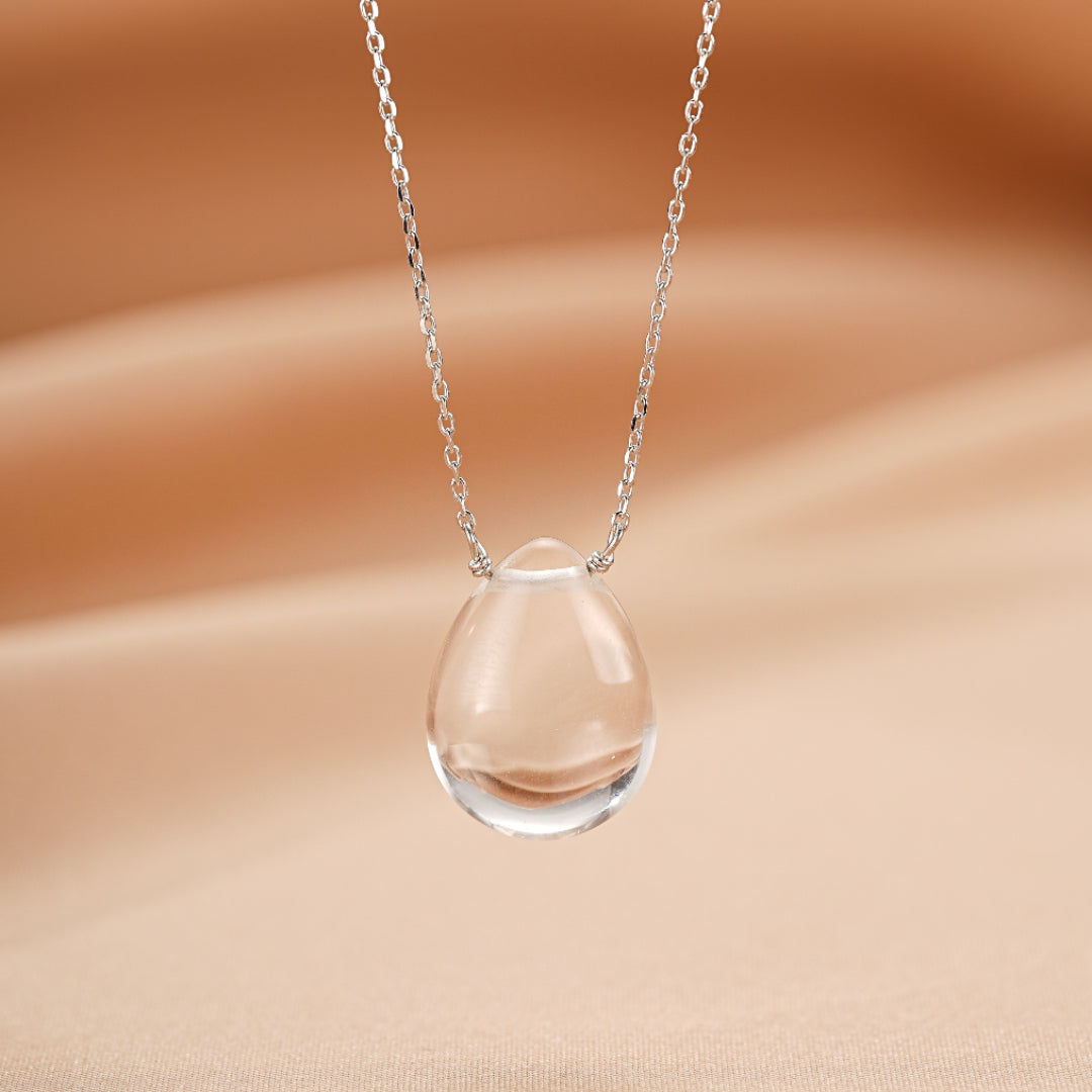 Goutte de cristal de roche transparente en forme de larme, suspendue à une fine chaîne en argent, sur un fond de soie beige.