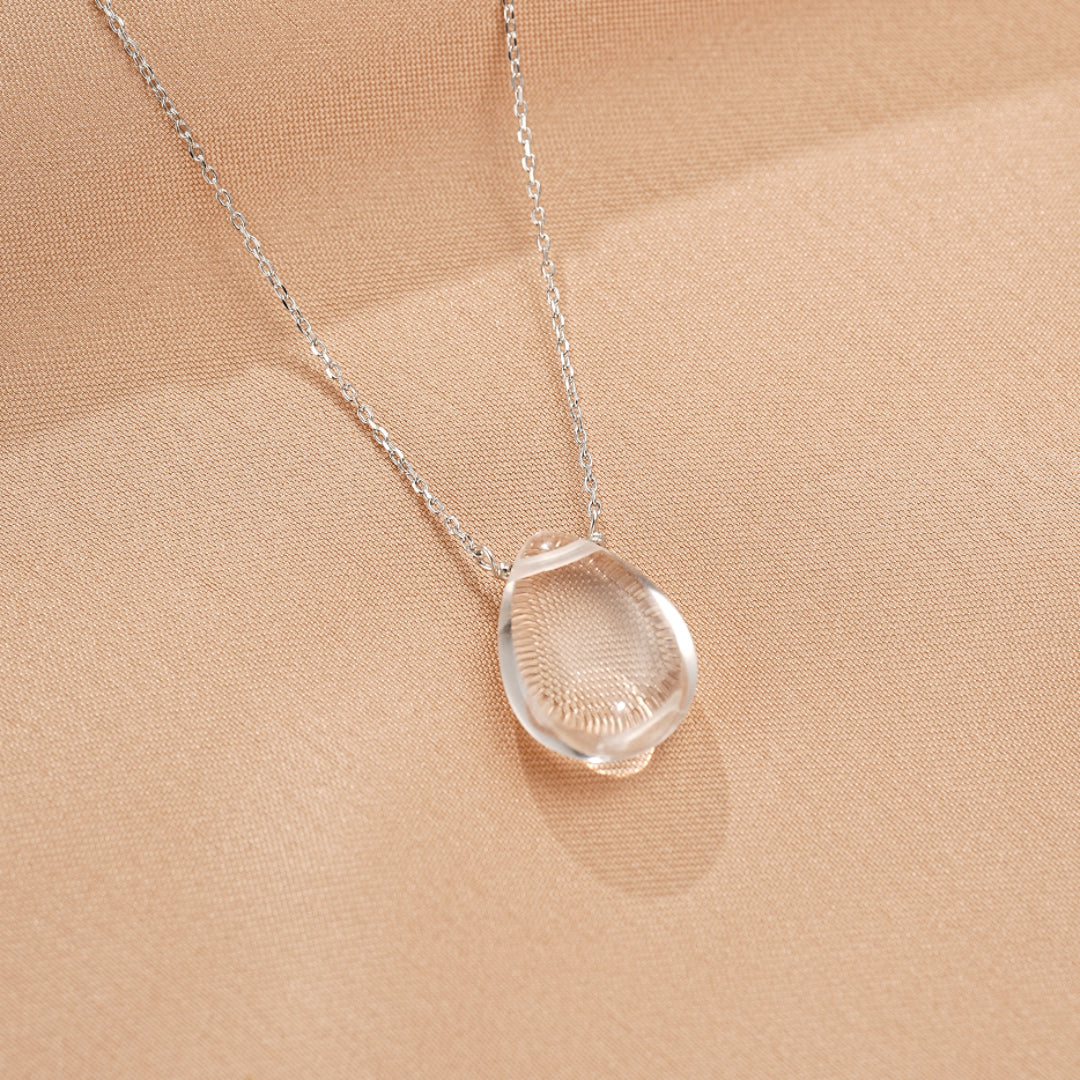 Gros plan sur un collier pendentif en forme de larme en cristal de roche clair, monté sur une fine chaîne en argent, posé sur un tissu beige soyeux.