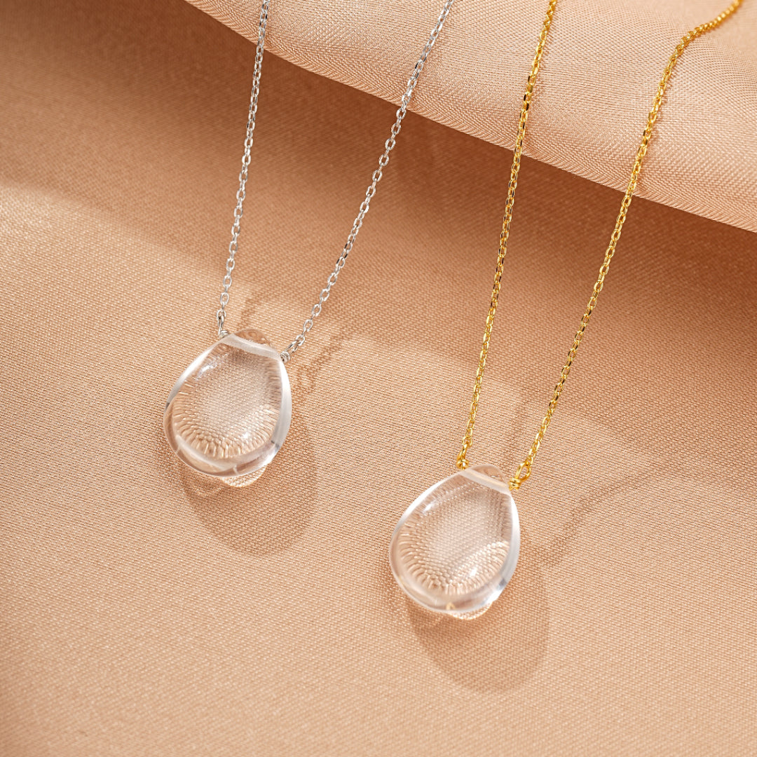 Deux colliers délicats avec des pendentifs en forme de larme en cristal de roche transparent sur des chaînes argentées et dorées, posés sur un fond beige texturé.