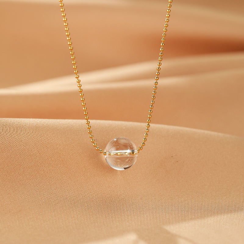 Collier en or avec une sphère en cristal de roche transparente au centre, sur fond de tissu beige drapé.