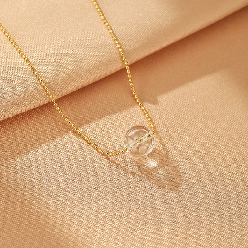 Collier en or avec une sphère en cristal de roche transparente au centre, sur un fond texturé beige.
