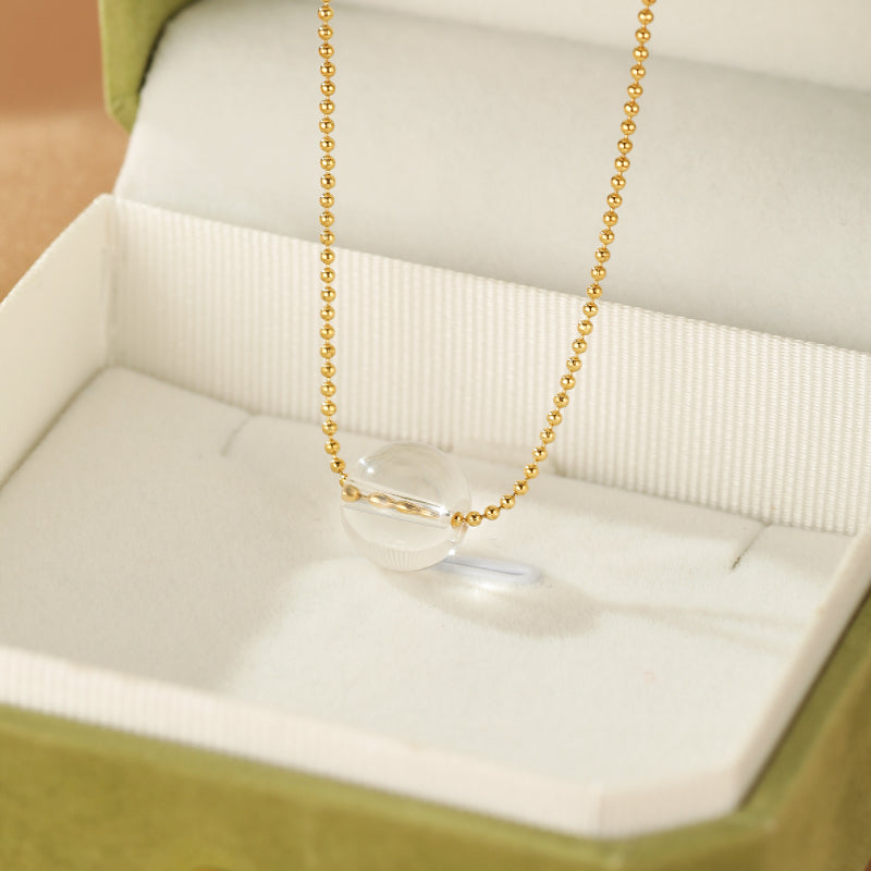 Collier en plaqué or avec une sphère de cristal de roche transparente et facettée, présenté dans un écrin vert.