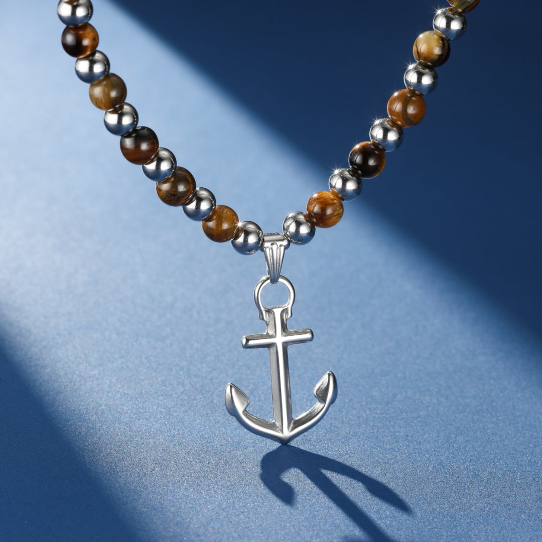 Collier pour homme avec un pendentif ancre marine en argent, entouré de perles œil de tigre et d'acier inoxydable sur fond bleu.