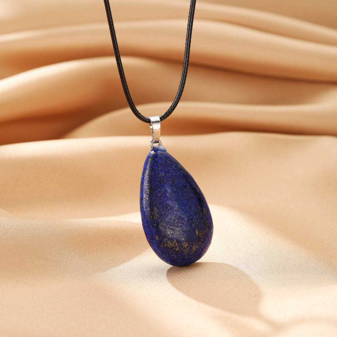 Collier pendentif avec une pierre de lapis-lazuli bleue en forme de goutte, suspendu à un cordon noir sur un fond satiné beige.