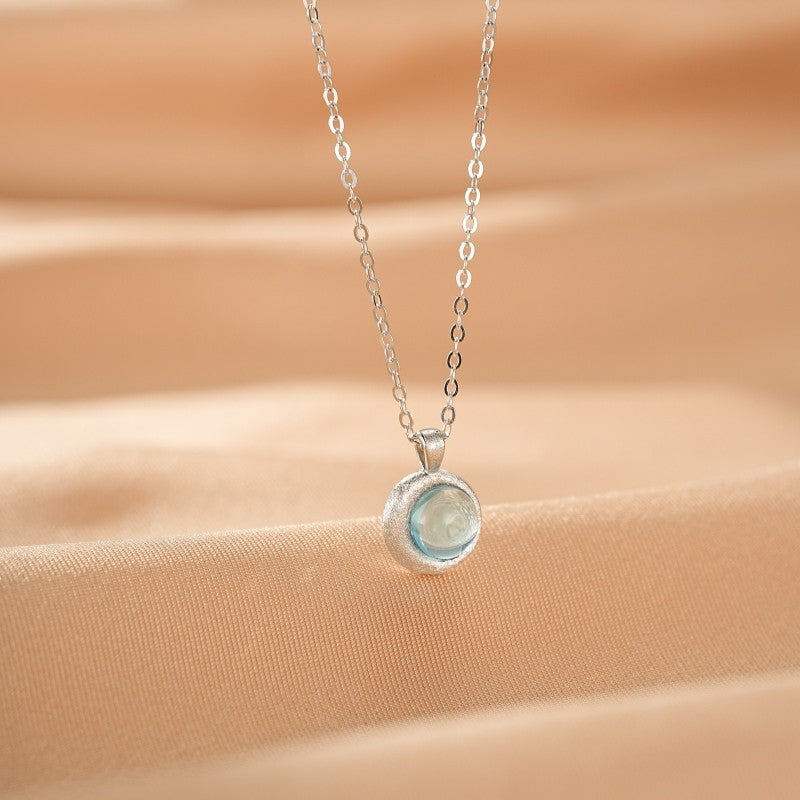 Collier en argent avec un pendentif rond en aigue-marine rappelant la lune, sur un fond beige satiné.