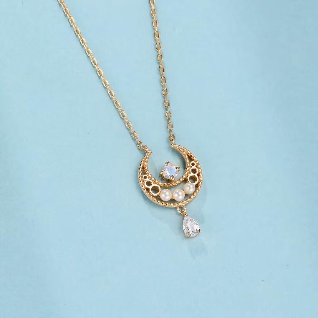 Collier en argent plaqué or en forme de croissant de lune orné d'une pierre de lune carrée, de petites perles et d'une goutte en cristal sur fond bleu clair.