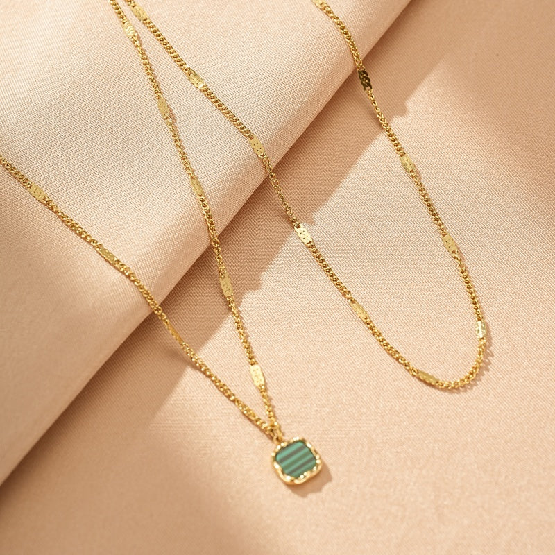 Collier doré avec un pendentif carré en malachite verte aux rayures distinctes, sur un fond de tissu beige.