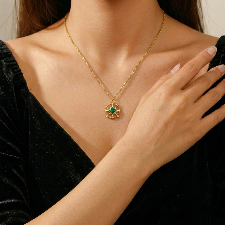 Collier en or avec un pendentif carré vert en malachite orné de détails dorés rappelant une boussole ou une étoile, porté par une femme sur un vêtement noir.
