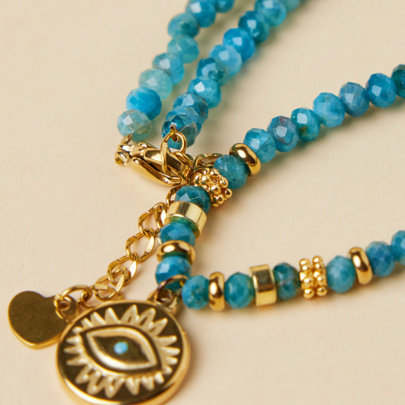 Gros plan sur un collier en apatite bleue avec un pendentif œil turc doré et des perles dorées, mettant en valeur le fermoir et la chaîne.