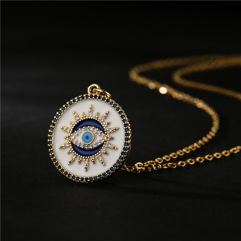 Gros plan sur un pendentif de collier œil du mal de forme ronde, orné d'un œil bleu central, entouré d'une étoile dorée sur fond blanc, le tout bordé de zirconiums bleus et dorés. La chaîne du collier est en or.