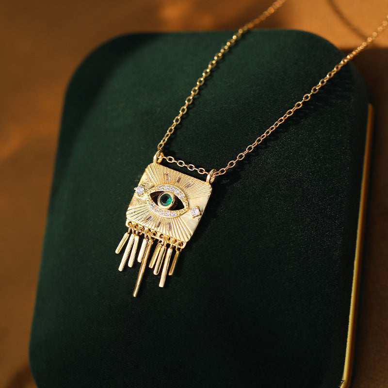 Collier plaqué or avec un pendentif carré texturé orné d'un œil stylisé avec une pierre verte au centre, entouré de petits cristaux et de rayons, avec des franges tombantes en bas. La chaîne est fine et dorée.