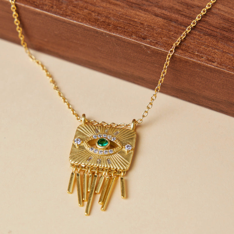 Collier en argent plaqué or avec un pendentif carré orné d'un œil stylisé vert et blanc, entouré de cristaux, et de franges dorées pendantes.