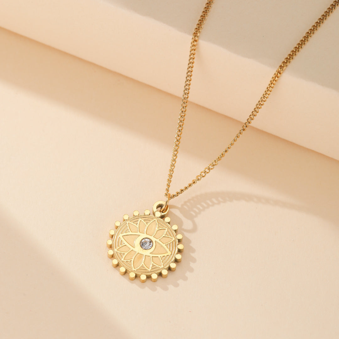 Collier en or avec pendentif rond gravé d'un œil stylisé et de rayons de soleil, orné d'une petite pierre au centre.