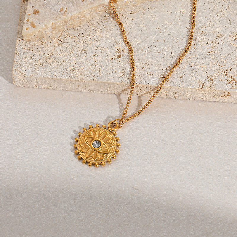 Gros plan sur un collier en or avec un pendentif rond orné d'un mauvais œil stylisé et d'une petite pierre bleue au centre, sur un fond clair et texturé.