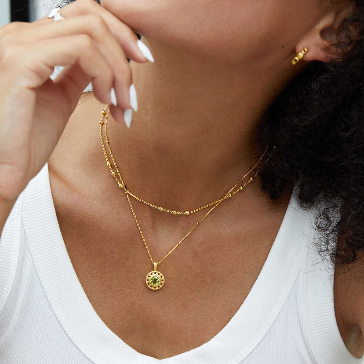 Mannequin portant un collier superposé en argent plaqué or, avec un pendentif rond serti d'une pierre verte péridot au centre, et un collier plus court orné de petites perles dorées.