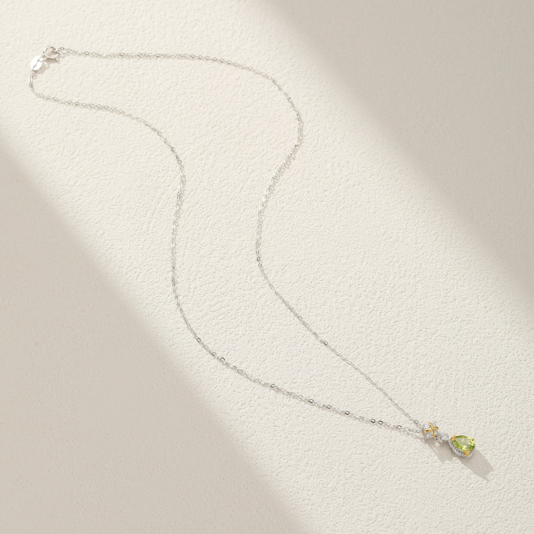 Collier en argent avec pendentif en forme de goutte de péridot et accents de petites pierres jaunes, vue de dessus sur fond texturé clair.