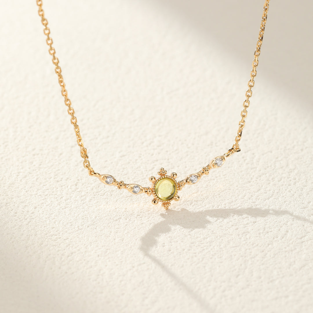Collier fin en argent plaqué or avec un pendentif central en péridot vert pâle entouré de petits cristaux et de motifs ajourés.