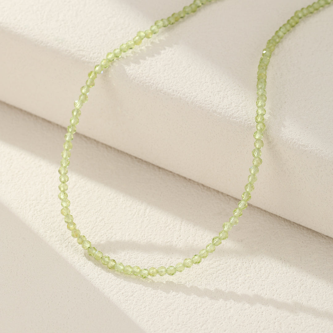 Collier ras du cou en perles de péridot facetées fines de couleur vert pâle, posé sur un fond blanc texturé.