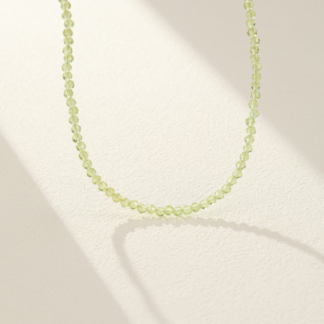 Collier ras-du-cou délicat fait de petites perles rondes de péridot facettées vert clair, suspendu sur un fond blanc avec une ombre douce projetée.