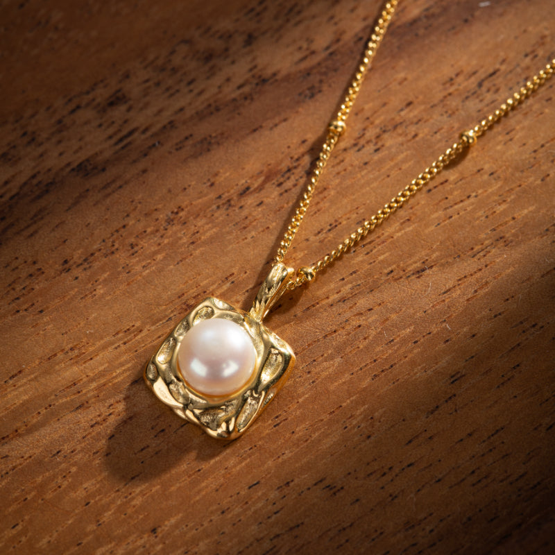 Pendentif carré texturé en argent plaqué or avec une perle rose pâle au centre, sur fond de bois sombre.