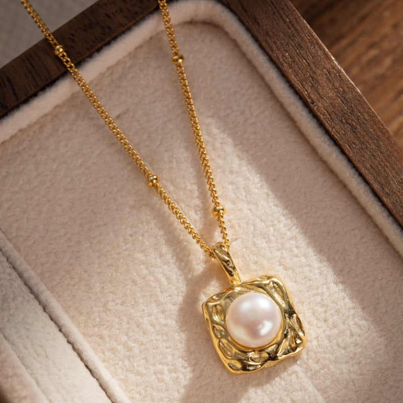 Collier en argent plaqué or avec un pendentif carré texturé retenant une perle blanche nacrée. La chaîne est ornée de petites perles dorées.