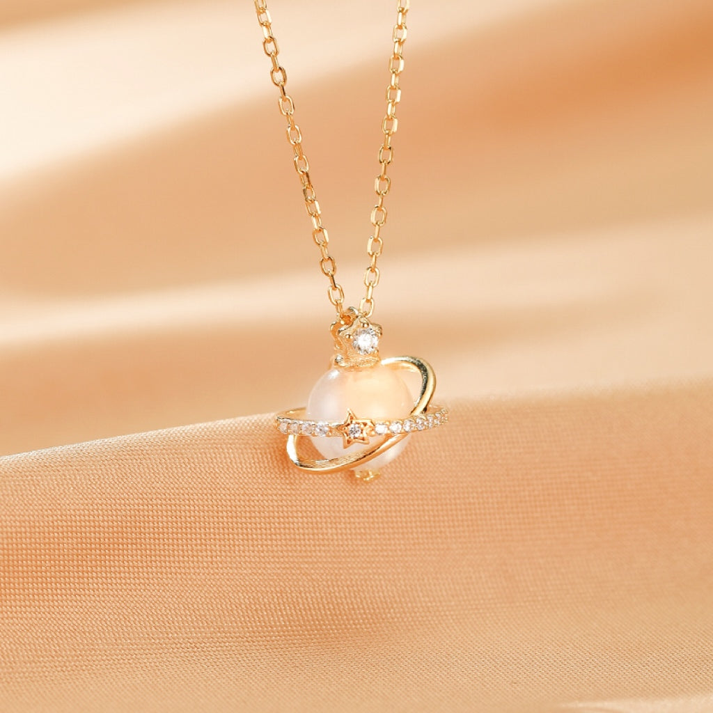 Collier "Oorbite Céleste" en argent plaqué or avec une sphère nacrée, un anneau serti de cristaux et une étoile, le tout suspendu à une chaîne délicate.