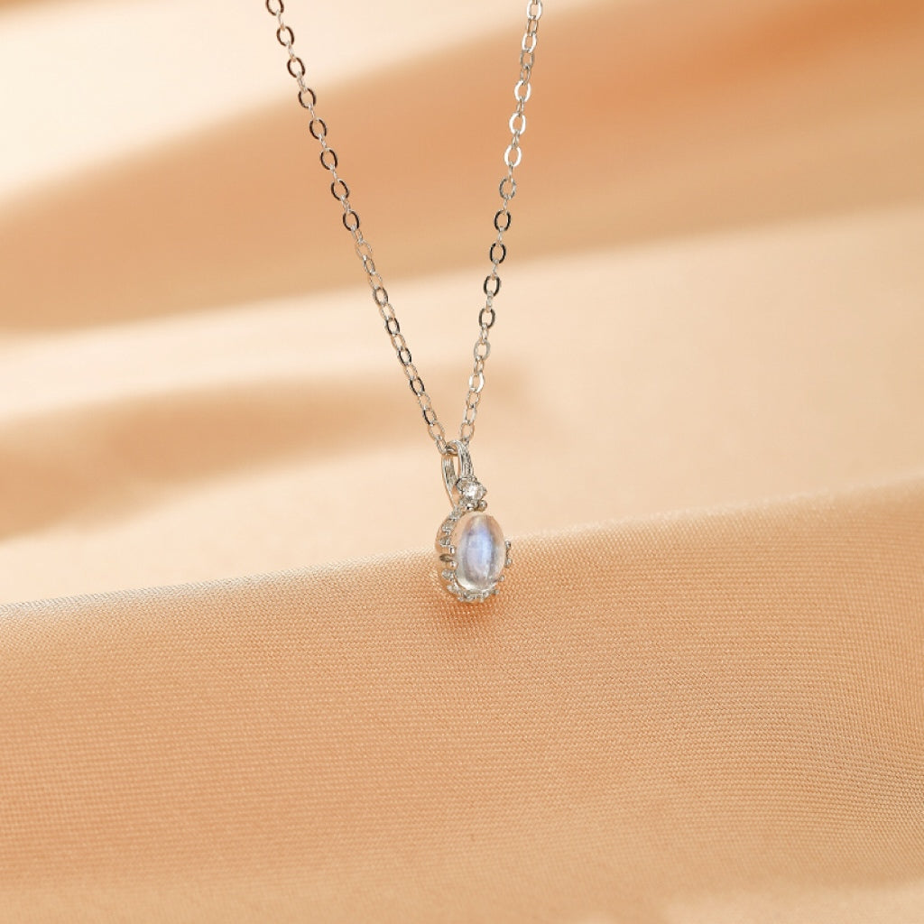 Collier "Rêve de Lune" avec pendentif ovale en pierre de lune bleue et chaîne fine en argent plaqué or, serti de petites pierres scintillantes, sur un fond satiné couleur pêche.
