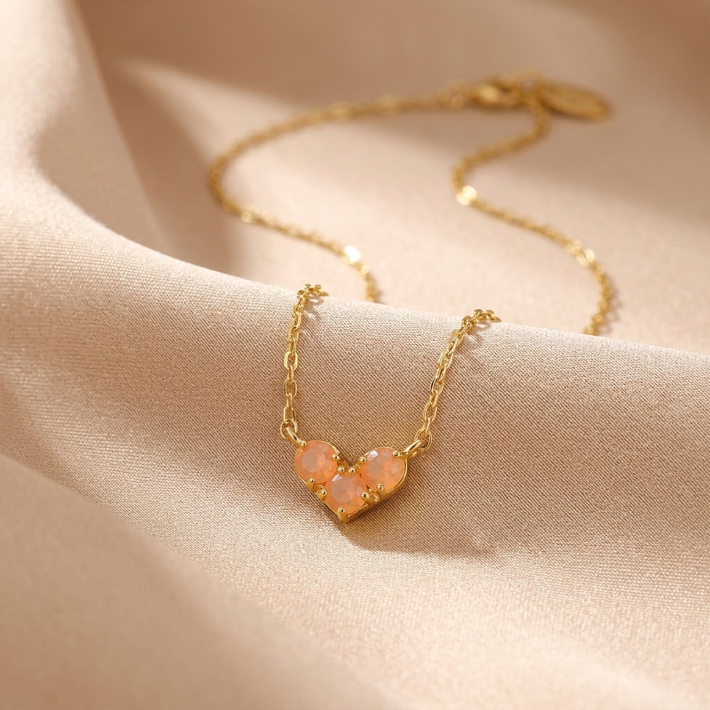 Collier en argent plaqué or avec pendentif en forme de cœur orné de trois pierres roses translucides, sur fond de tissu satiné couleur crème.