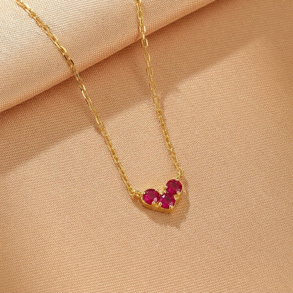 Collier en argent plaqué or avec trois petites pierres rouges disposées en forme de cœur sur un fond beige