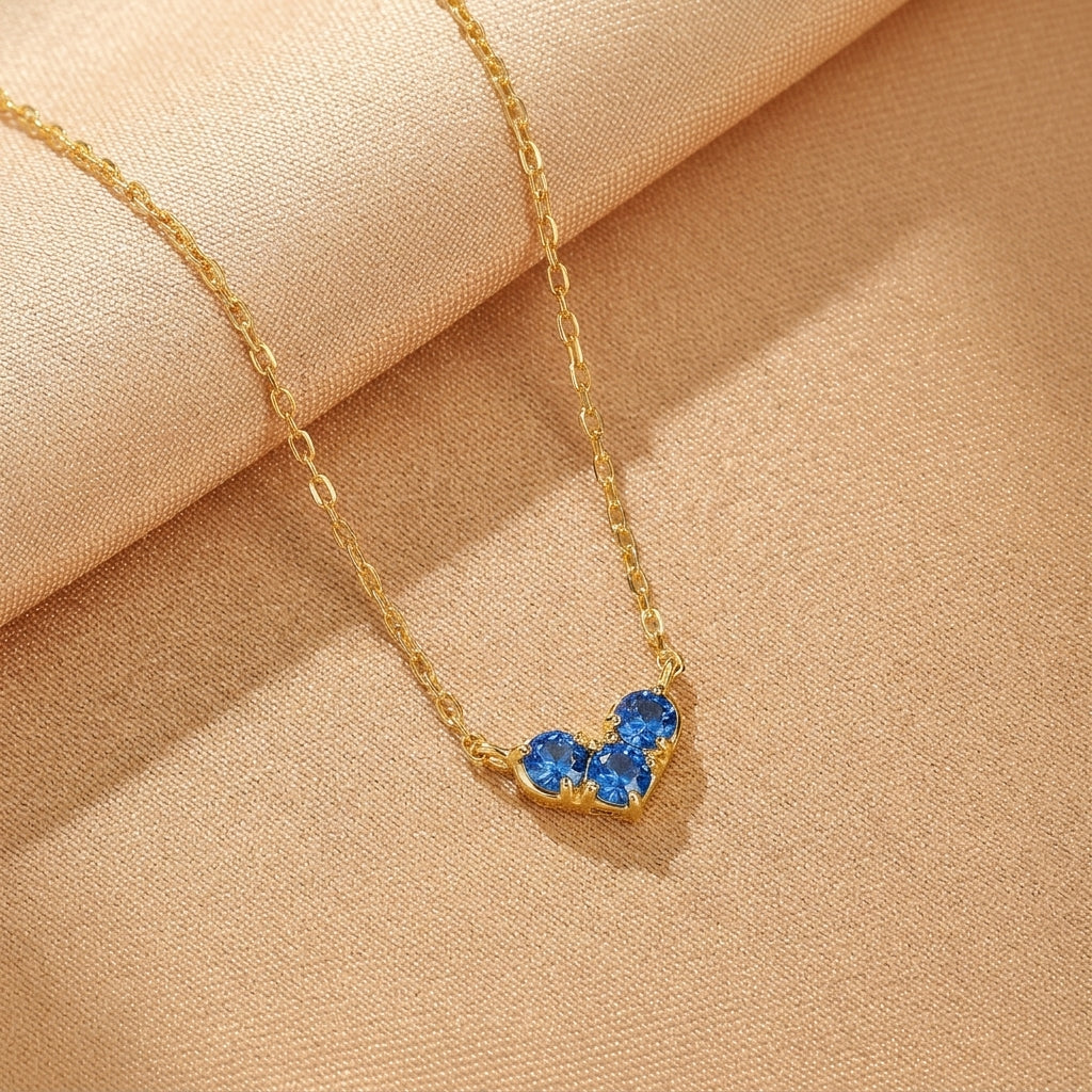 Collier en argent plaqué or avec trois pierres bleues en forme de cœur, sur un fond beige texturé.