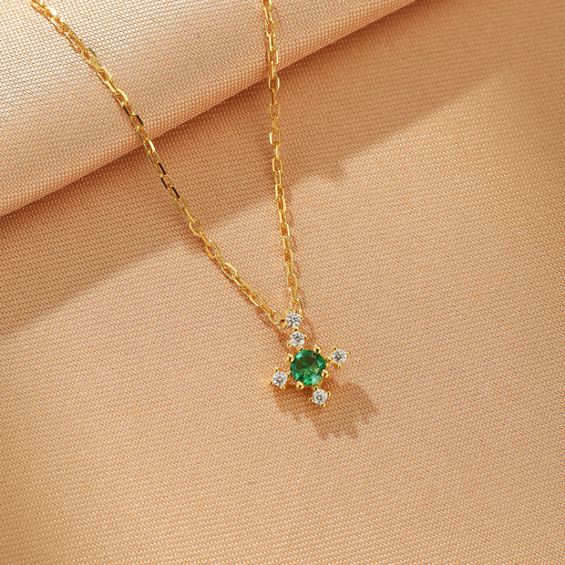 Gros plan sur un collier en argent plaqué or avec un pendentif en forme de croix orné d'une émeraude centrale et de quatre petits diamants de chaque côté, le tout sur un fond texturé beige.