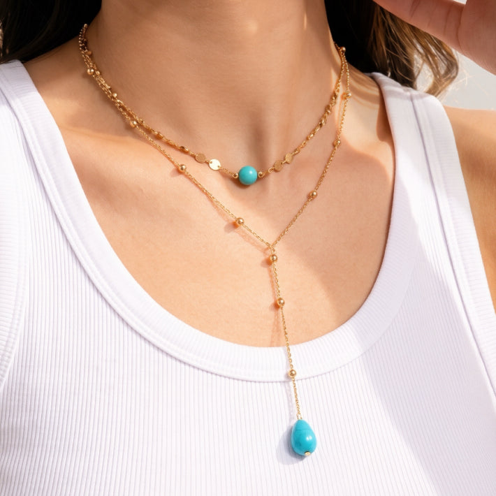 Un collier double chaîne plaqué or avec une perle turquoise ronde sur la chaîne supérieure et un pendentif turquoise en forme de goutte sur la chaîne inférieure.