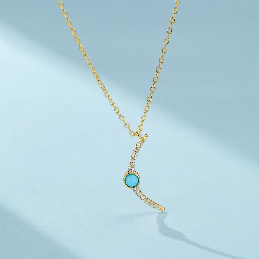 Collier en argent doré avec une pierre turquoise ronde et une courbe sertie de petits cristaux blancs, sur fond bleu clair