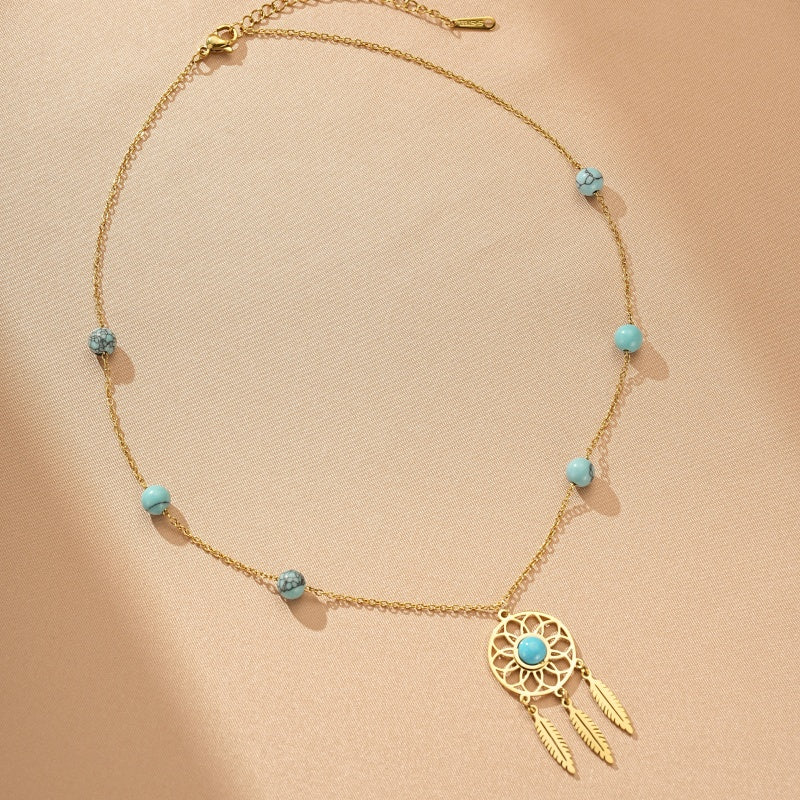 Collier doré avec un pendentif attrape-rêves orné d'une turquoise centrale et de trois plumes, agrémenté de perles bleues rappelant la turquoise le long de la chaîne.