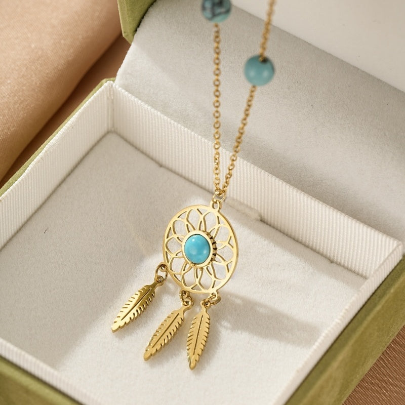 Collier doré avec pendentif attrape-rêves orné d'une turquoise centrale et de trois plumes, sur fond de boîte cadeau verte.