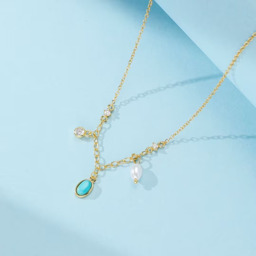 Collier en argent plaqué or avec une turquoise ovale et une perle, orné de petites pierres scintillantes, sur fond bleu ciel.