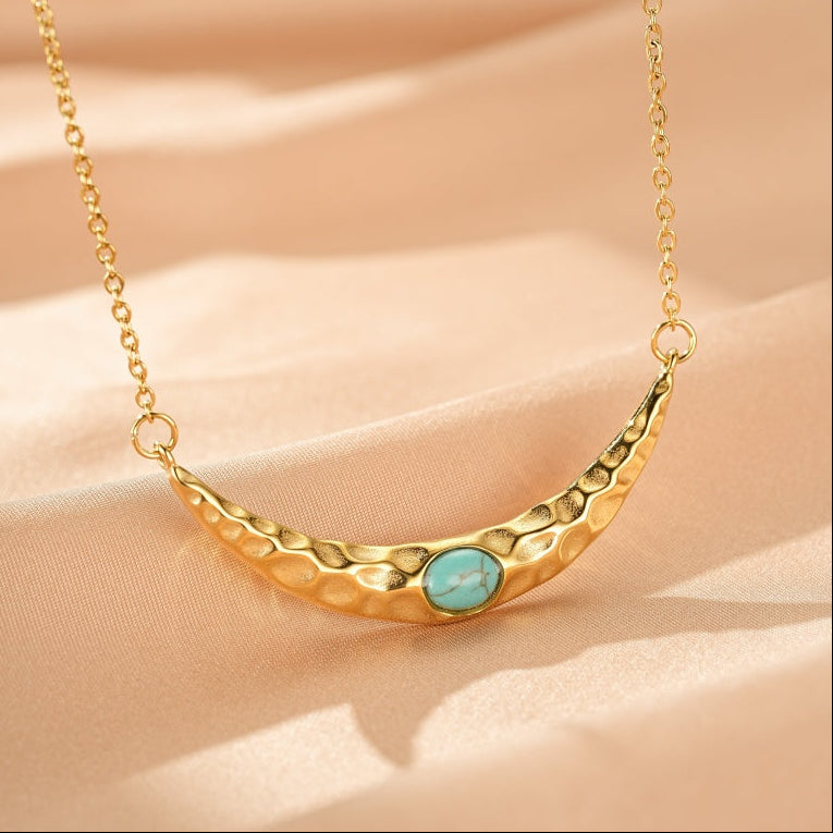 Collier en or texturé en forme de croissant de lune avec une pierre turquoise au centre, sur un fond de tissu beige