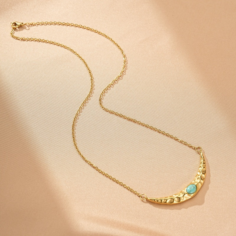 Collier en or avec pendentif croissant de lune martelé et une petite pierre turquoise au centre, sur un fond beige.