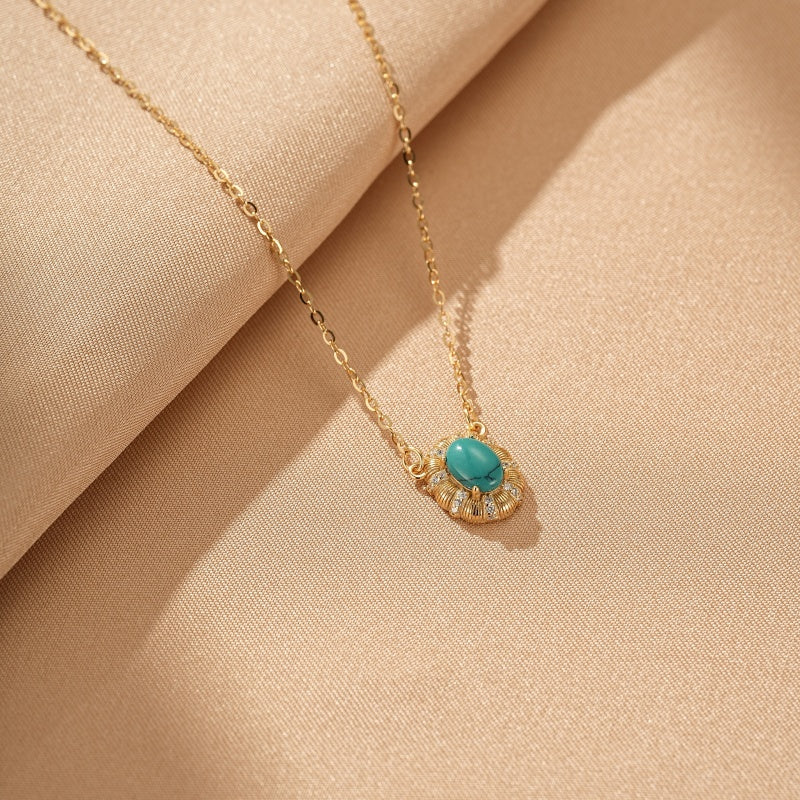Collier en argent plaqué or avec une pierre turquoise ovale entourée de petites pierres blanches scintillantes, présenté sur un tissu beige.