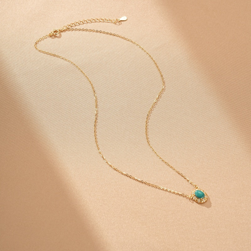 Collier fin en argent plaqué or avec une petite turquoise ovale au centre, sertie de minuscules cristaux brillants, sur un fond beige texturé.