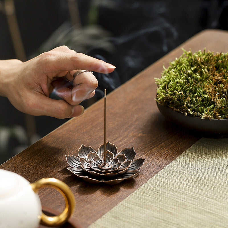 Encensoir "Lotus" en métal avec un bâton d'encens allumé et de la fumée s'élevant, sur un plateau en bois avec un petit bol de mousse verte et une théière en arrière-plan.