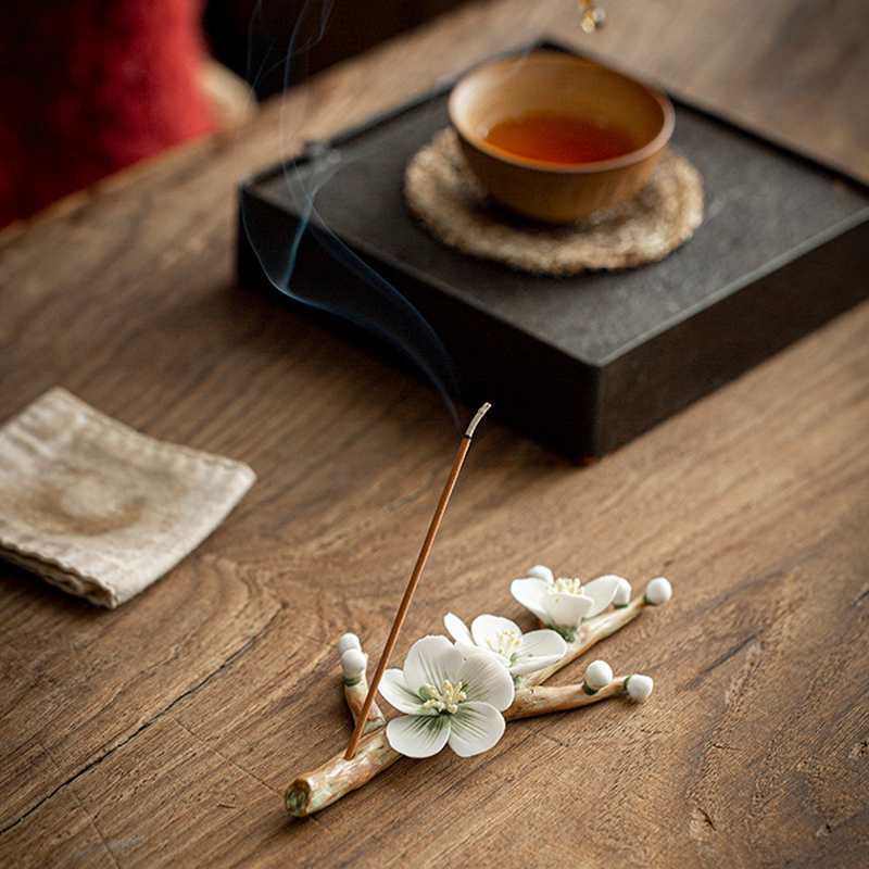 Encensoir en céramique fait main en forme de branche fleurie, avec un bâton d'encens allumé dégageant de la fumée, sur une table en bois.