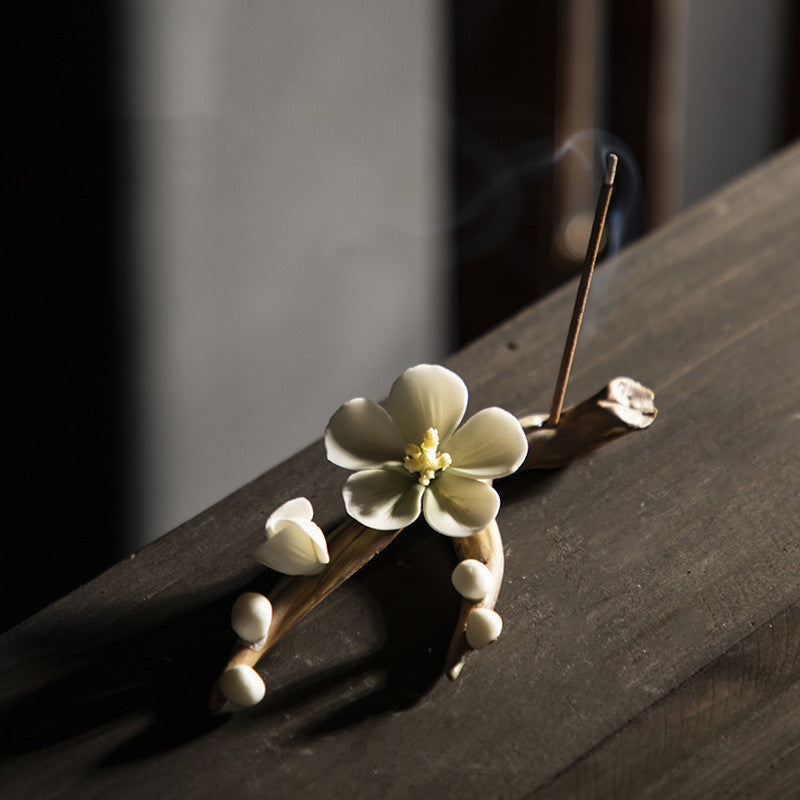 Encensoir zen artisanal en céramique avec une fleur blanche et des bourgeons, supportant un bâtonnet d'encens fumant.