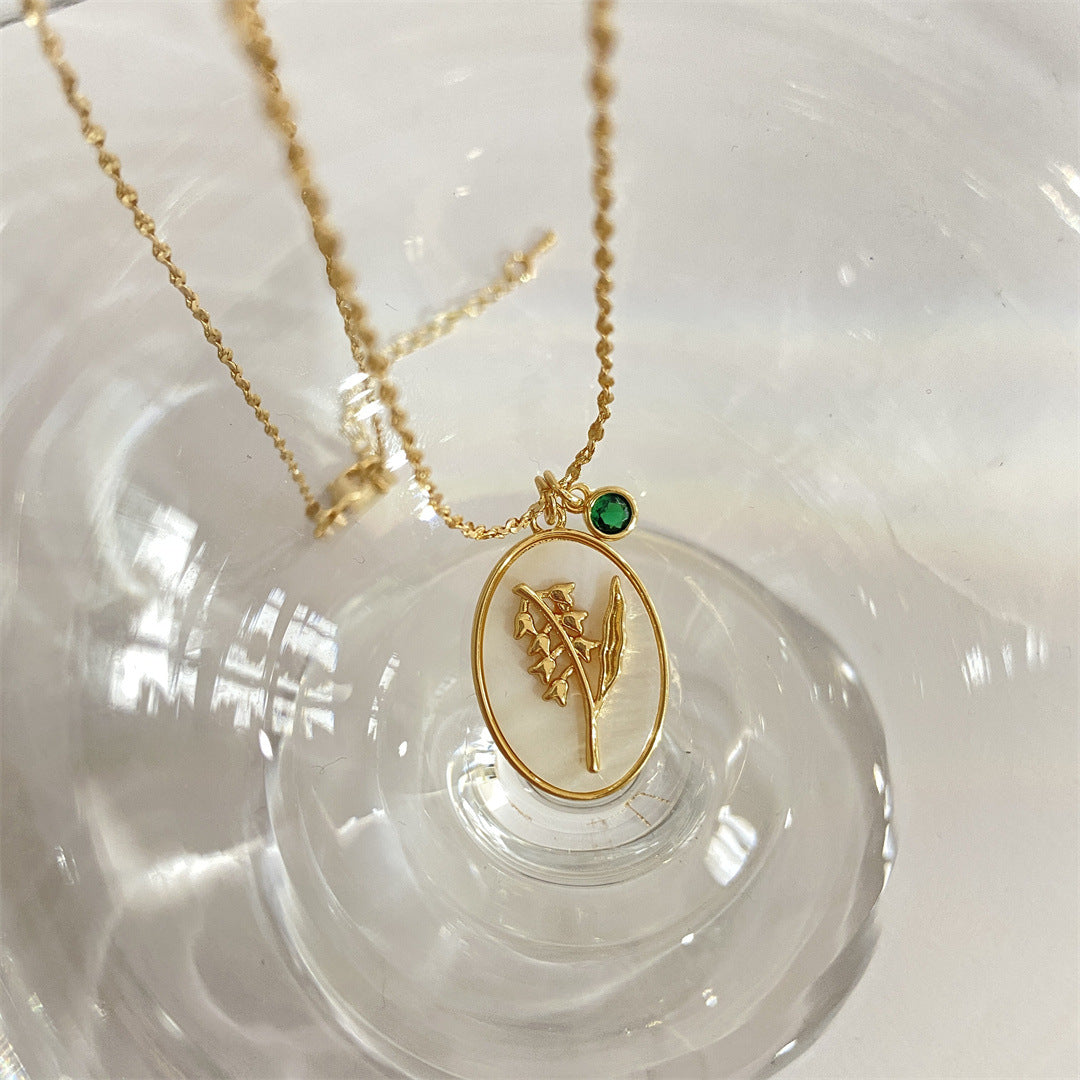 Pendentif en or avec un motif de muguet sur fond nacré ovale, une petite pierre verte à côté, chaîne fine.